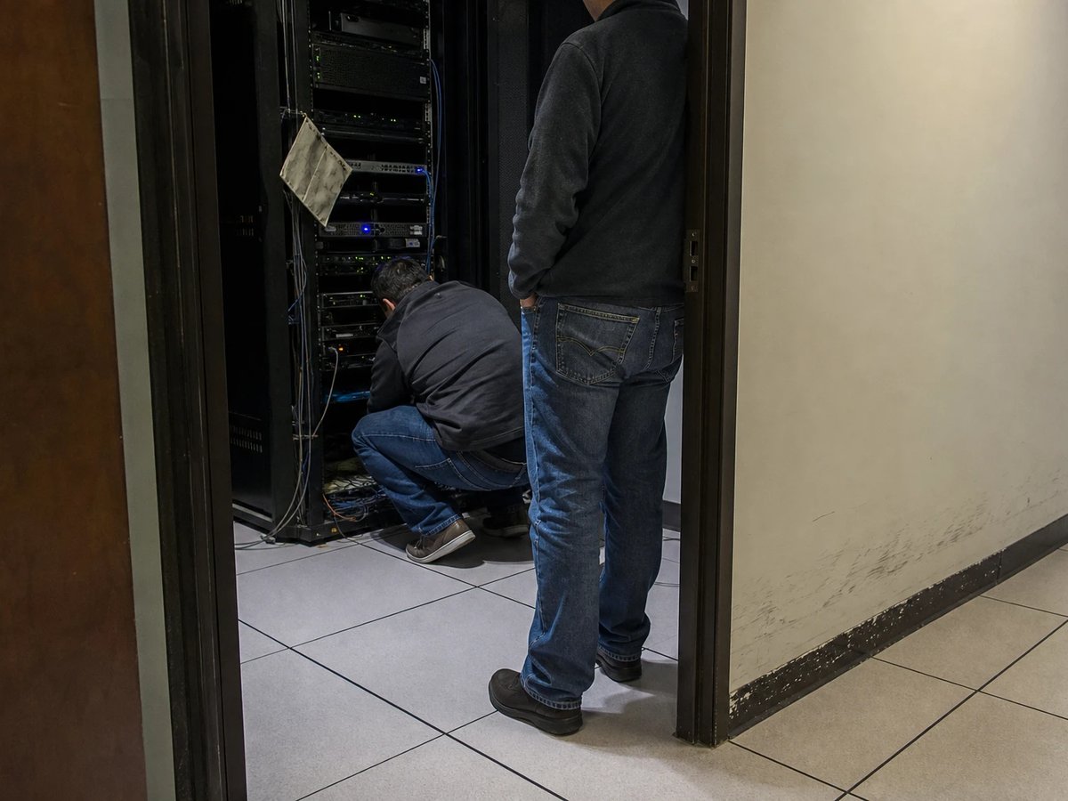 Zwei Plattform-Ingenieure in der offenen Tür eines kleinen Serverraums, einer prüft kauernd die Verkabelung am halb bestückten Rack