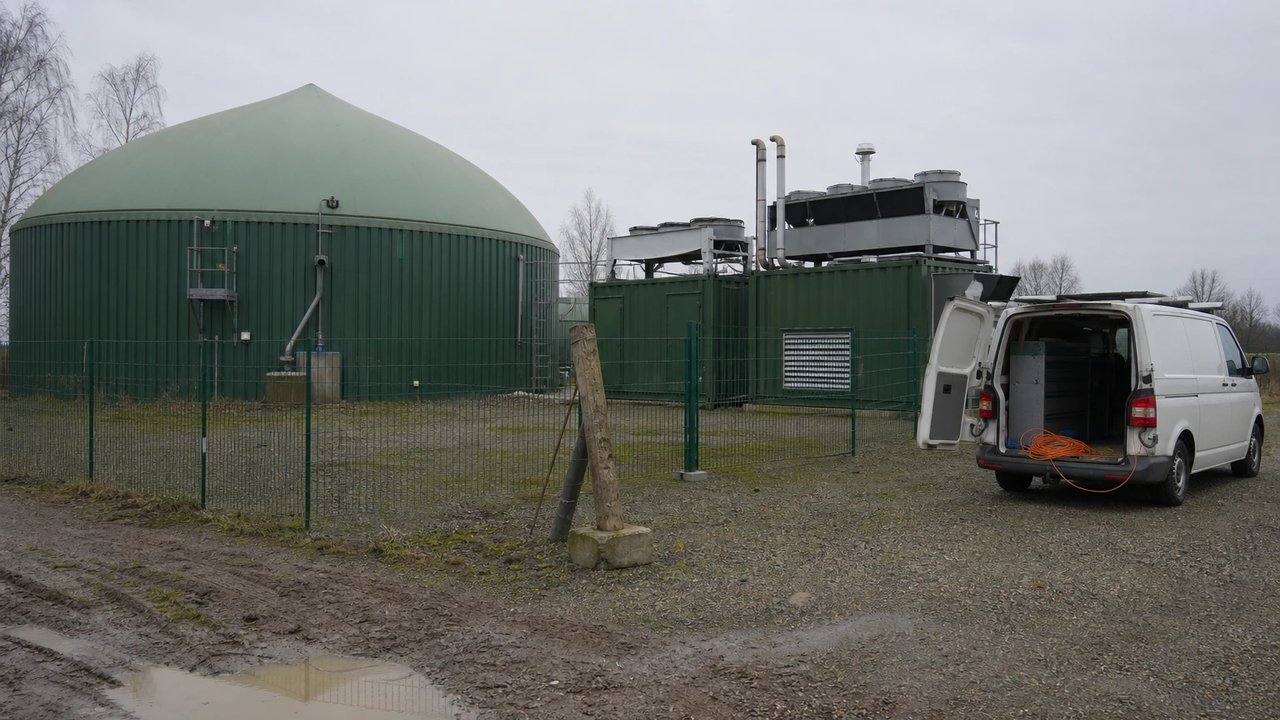 Biogasanlage mit grünem Fermenterdach und zwei BHKW-Containern am Dorfrand, davor ein Servicefahrzeug mit geöffneten Hecktüren.