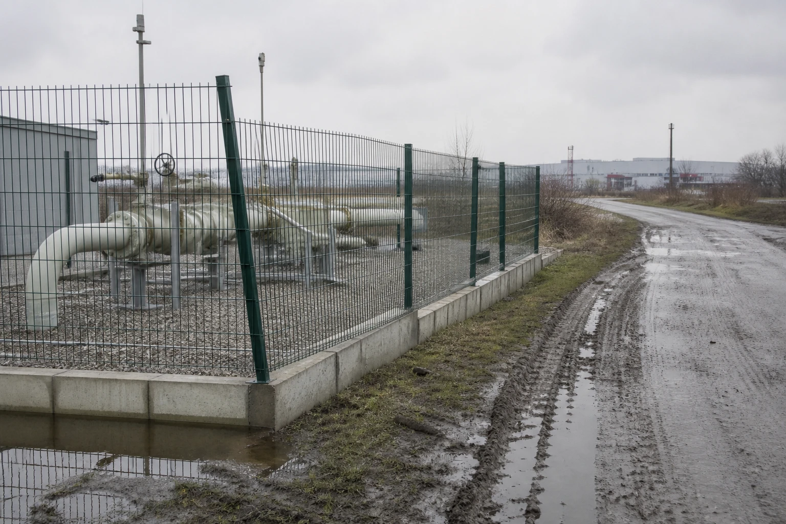 Gas-Druckregelstation hinter einem Maschendrahtzaun am Rand eines Industriegebiets, daneben ein Werksweg mit Reifenspuren im nassen Randstreifen