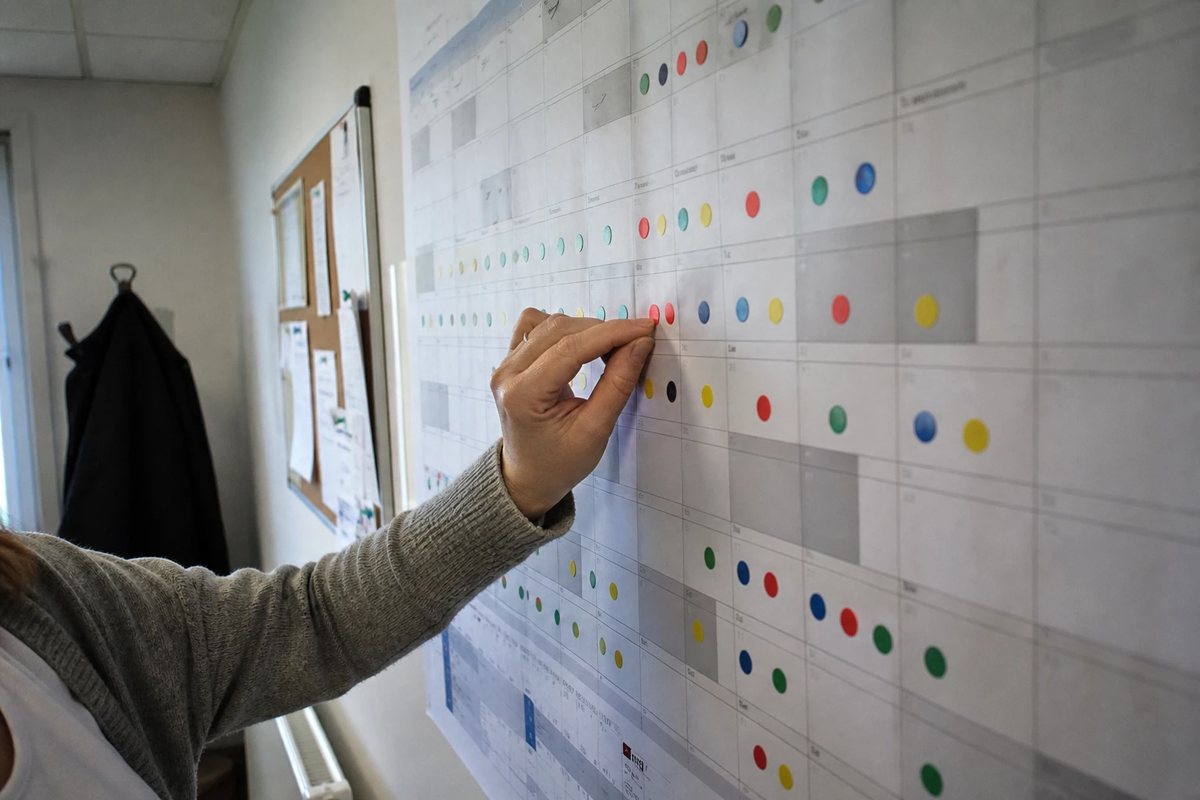 Wandkalender in einem Projektbüro: eine Hand setzt einen roten Klebepunkt auf einen Termin, Reihen farbiger Markierungen über die Monate