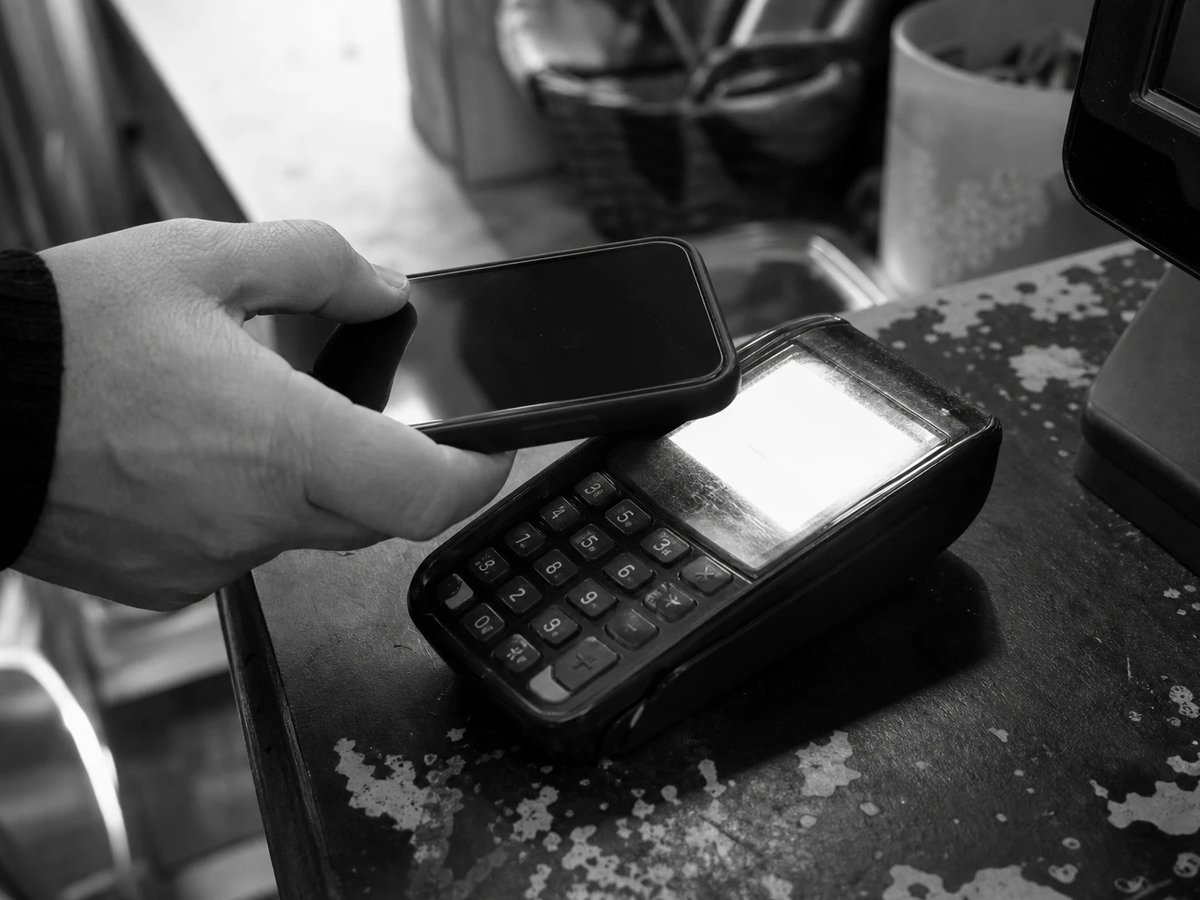 A hand holds a smartphone to a card terminal at a shop counter, a close-up of a contactless payment