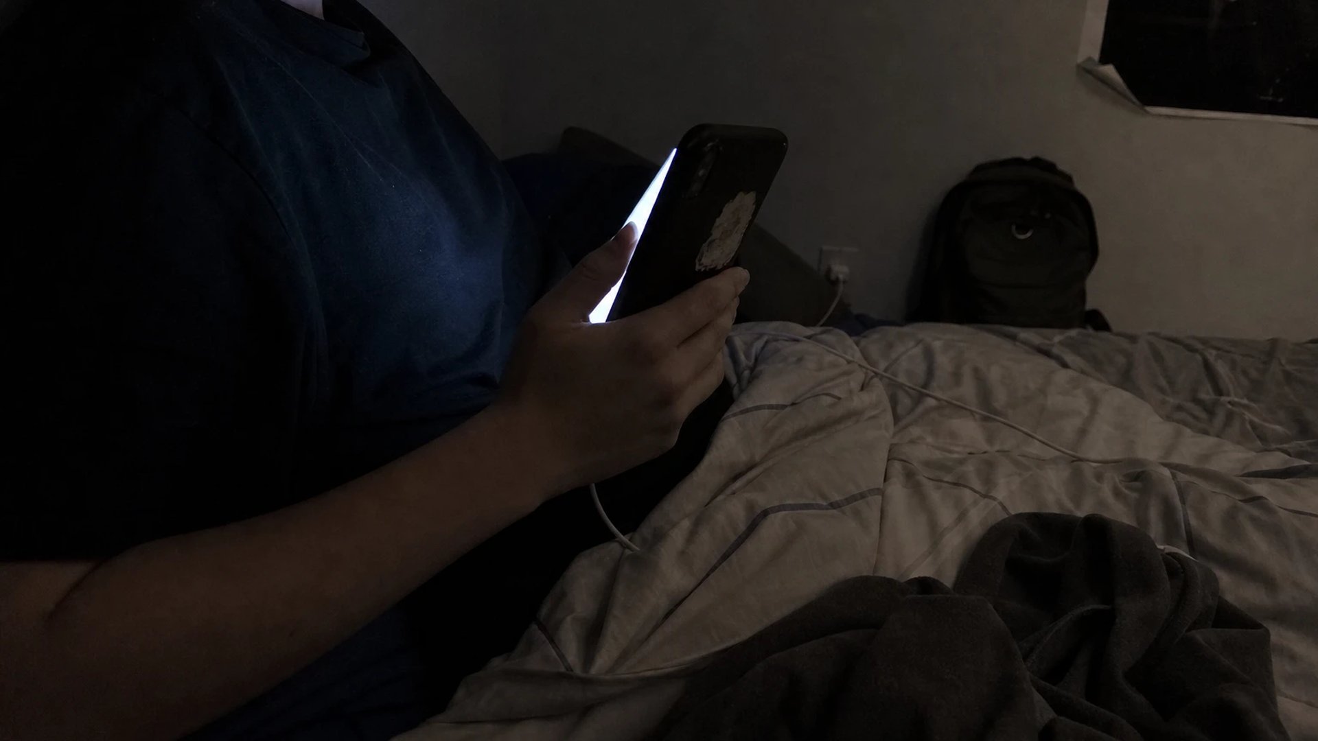 A teenager lies in a dark bedroom at night looking at a glowing smartphone screen, the face kept in shadow