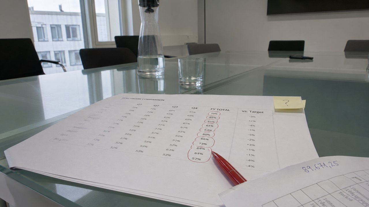 Printed benchmark comparison chart on a conference table with red pen circling a percentage column, in a German office meeting room