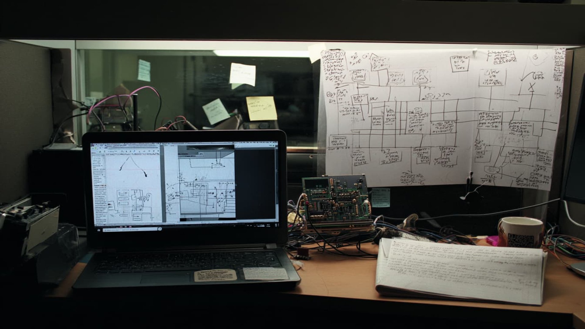 Worn workstation of an AI engineer with laptop, circuit boards, and handwritten diagrams on the wall
