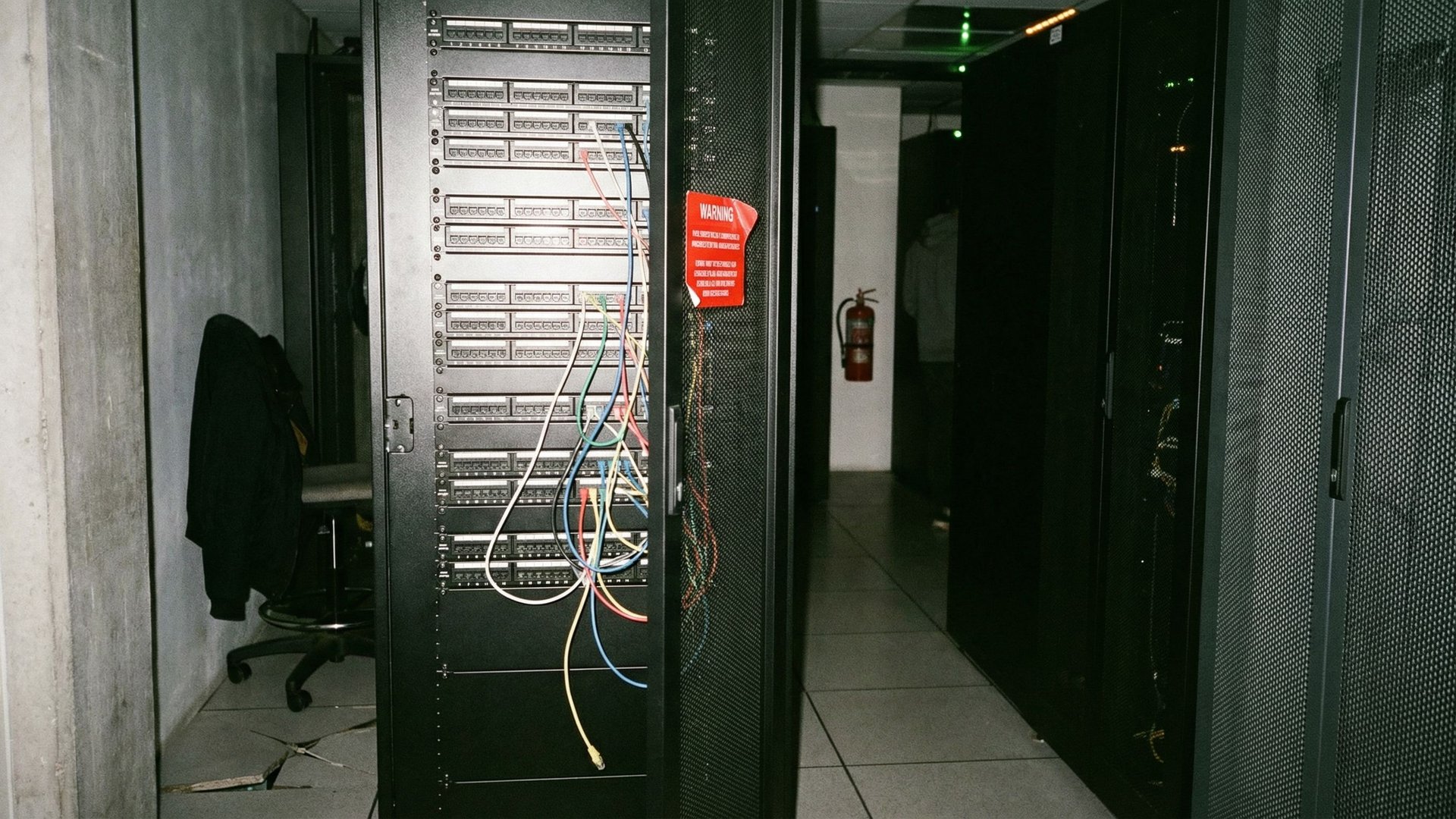 Server rack patch panels in a colocation corridor with loose ethernet cables and warning stickers, captured with flash photography