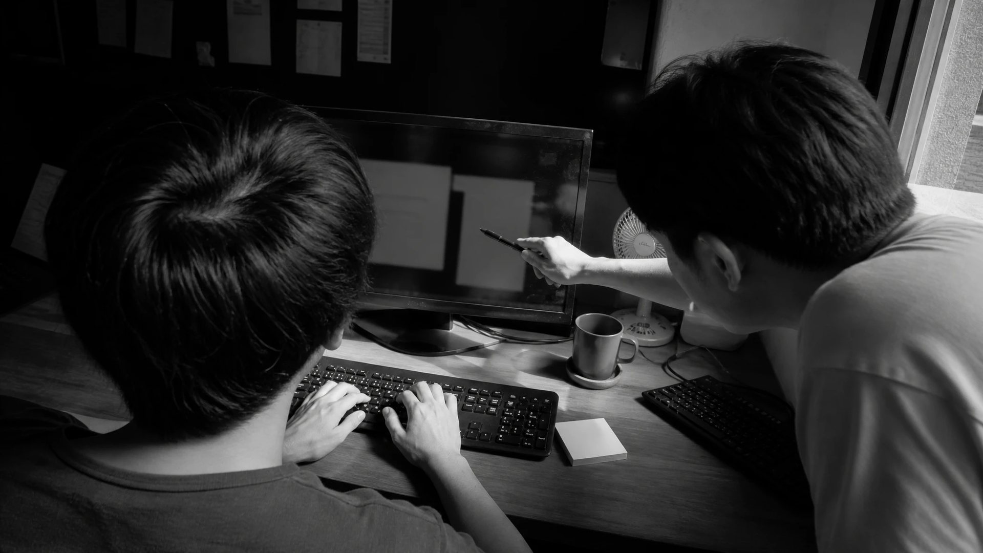 Two developers at a desk photographed from behind, one pointing a pen at the monitor