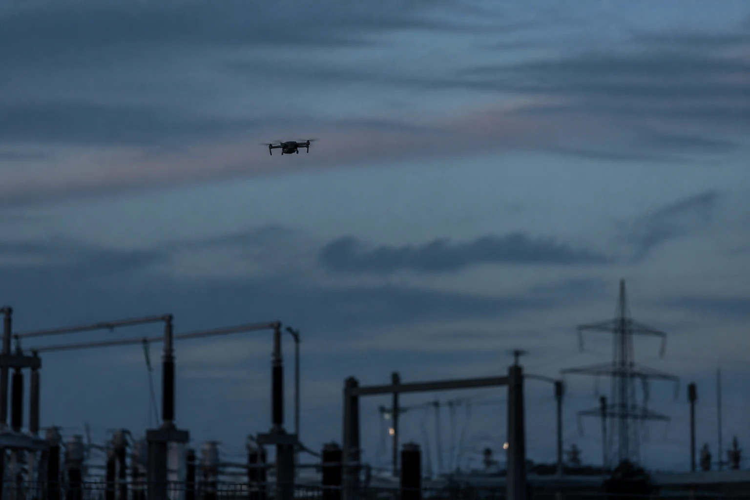 small commercial quadcopter drone as a dark silhouette at dusk above the equipment of an electrical substation
