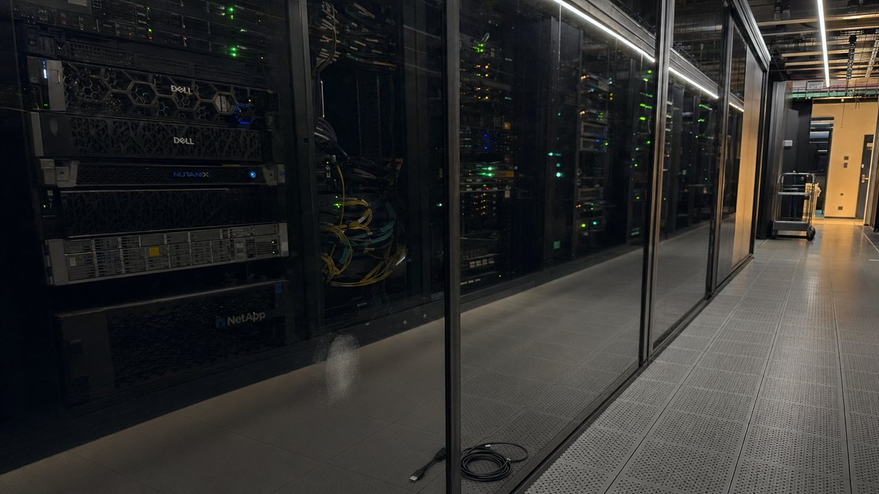 Long row of server racks behind a glass partition in a European colocation data hall, patch cable bundles and small green and amber status LEDs visible, a maintenance trolley parked at the far end of the corridor