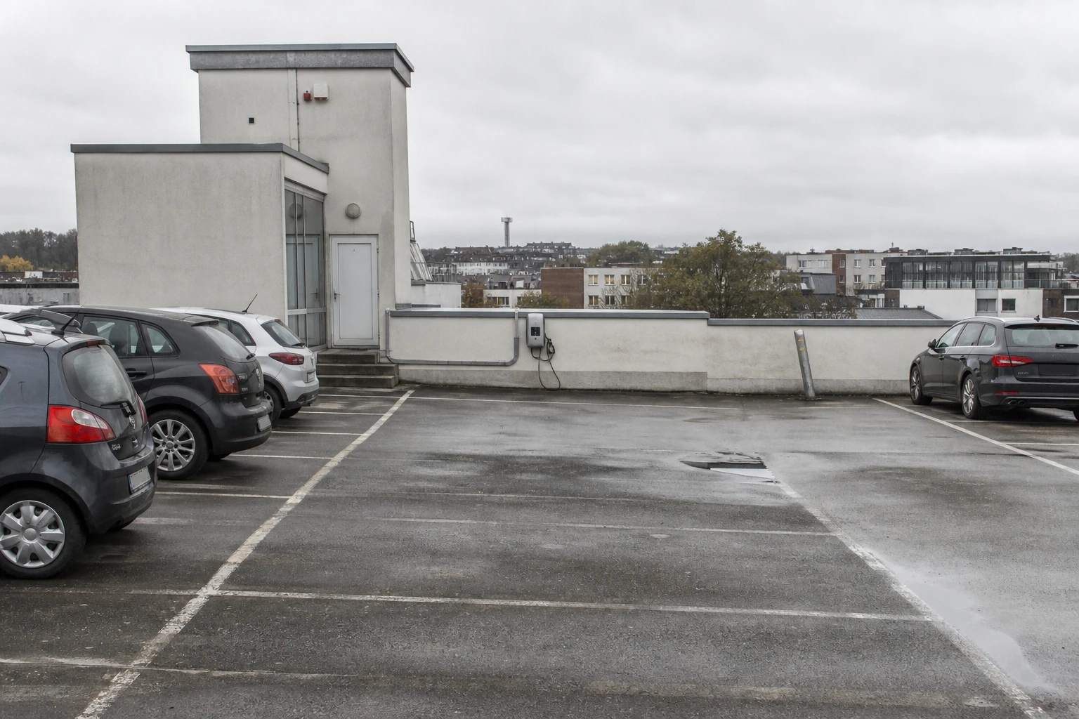 Open-air rooftop parking deck of a commercial building with parked cars, a single wall-mounted charging unit at the end of a row and a cable duct, low-rise commercial buildings beyond under a grey sky