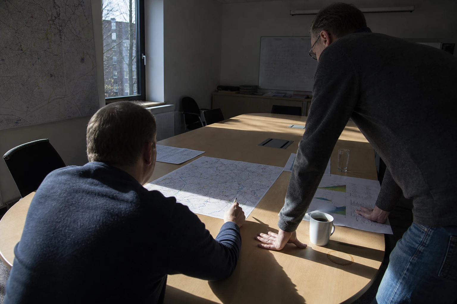 Two network planners at a meeting table comparing a printed medium-voltage plan with further documents, window light falling at an angle across the table