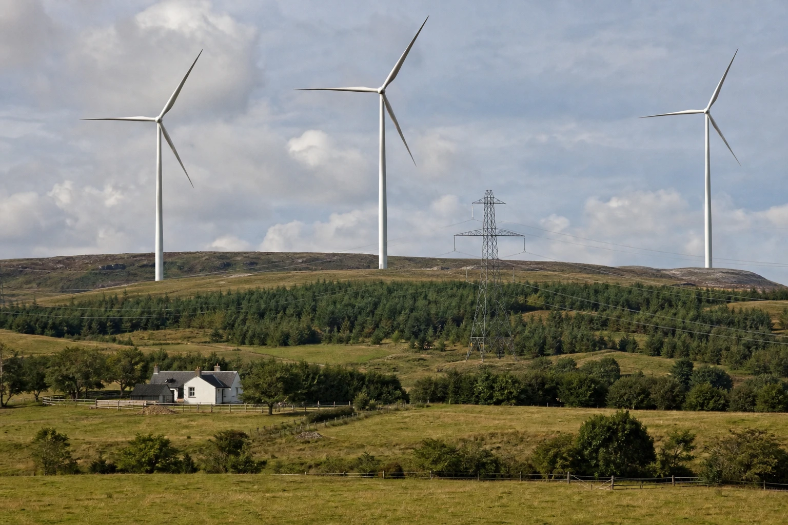 Several wind turbines on green hills with fields, a farmhouse and a power line, a cloudy sky
