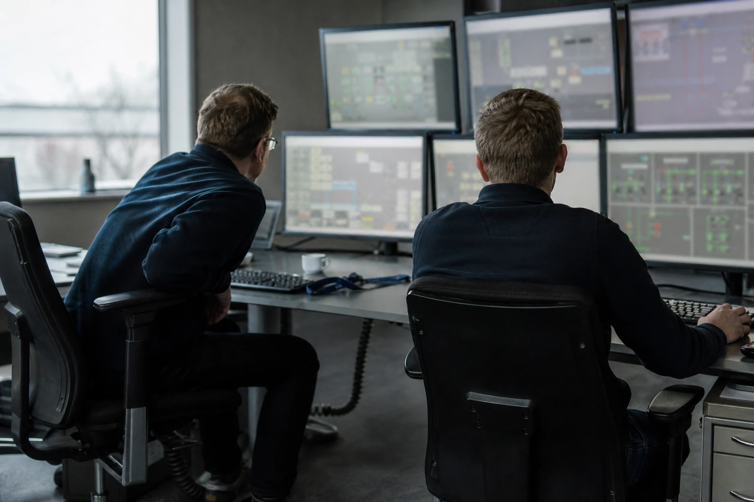 two engineers in the control room of a transmission system operator watching grid schematics and charts on several monitors