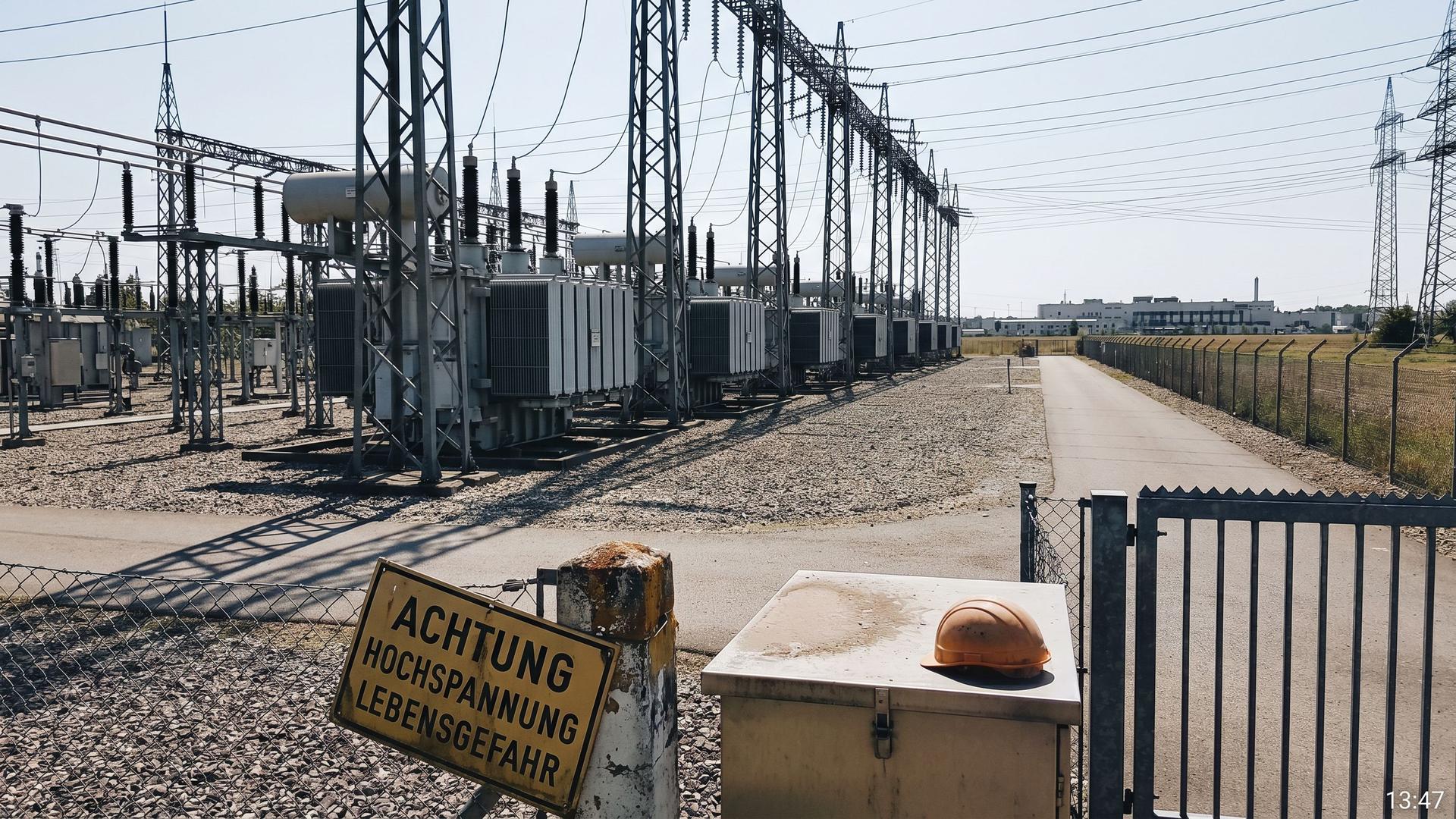 Electrical substation with transformers and steel lattice pylons in midday light with a distant data center on the horizon, symbol for the physical interface between AI infrastructure and the transmission grid