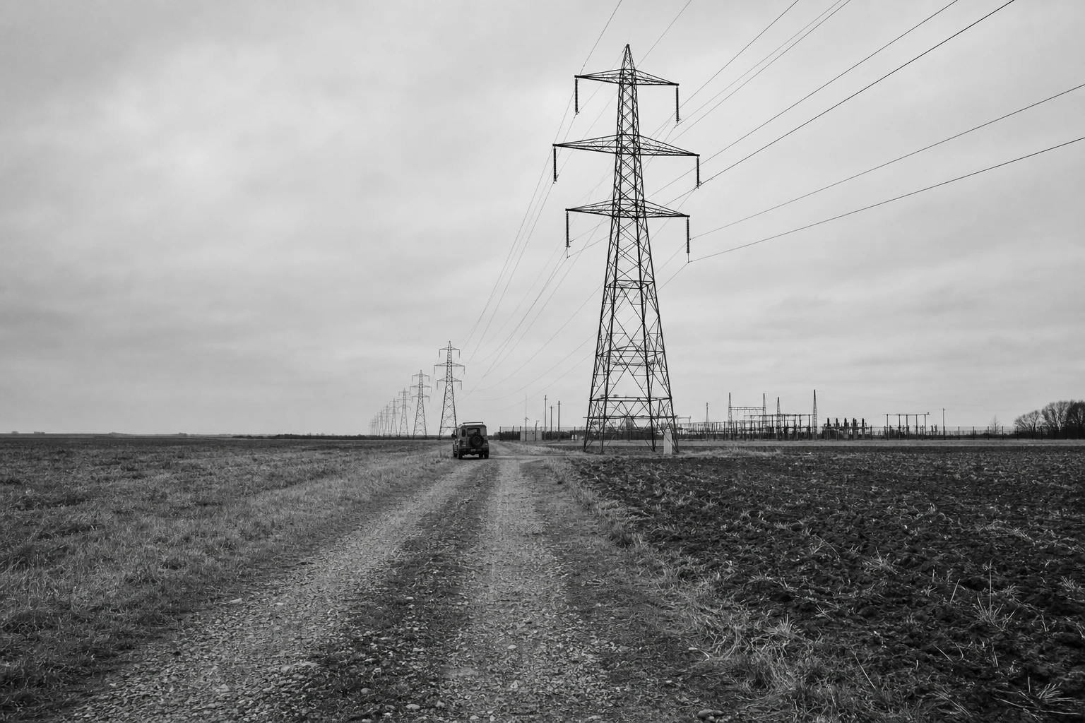 High-voltage transmission pylons across farmland under an overcast sky, a maintenance track leading to the nearest pylon