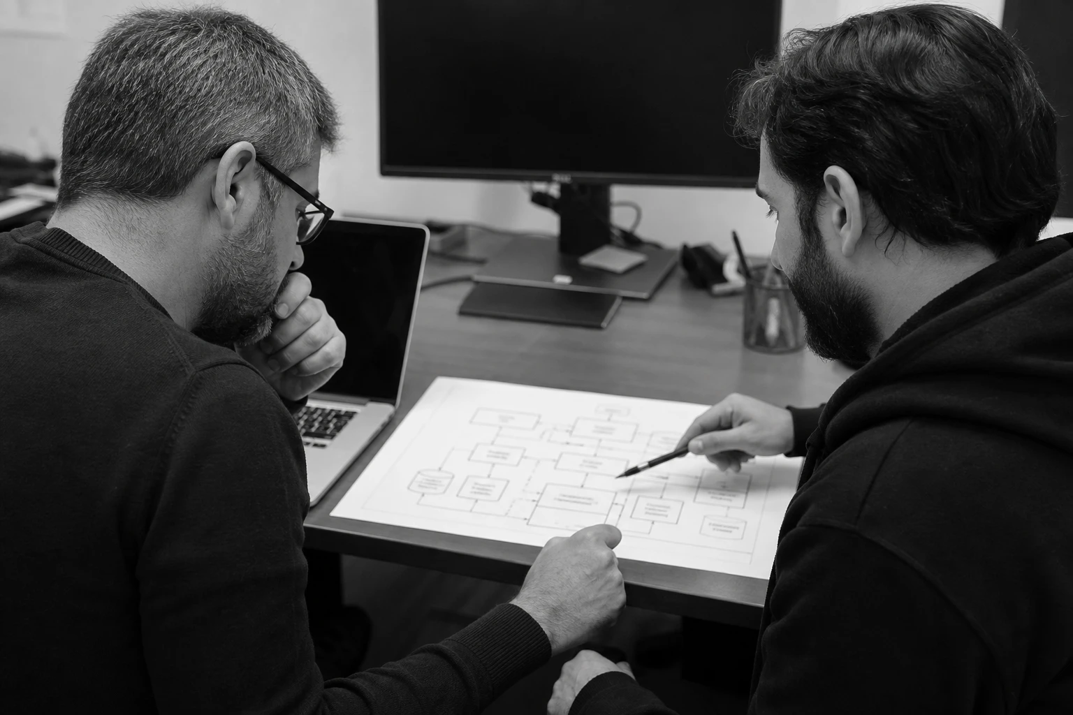 Two IT managers at a desk reviewing a printed system architecture diagram, black and white
